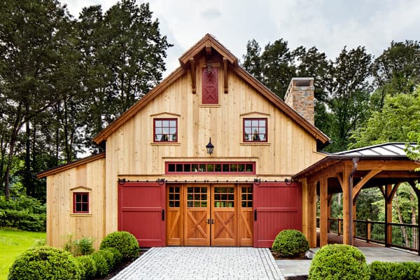 A rustic wooden party barn with red accents, large double doors, and multiple windows, surrounded by green bushes and trees, under a partly cloudy sky.