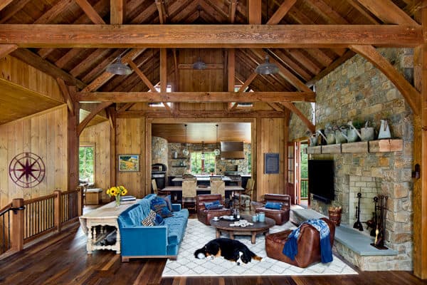 Rustic living room with wooden beams, stone fireplace, and cozy furnishings. Blue sofa, brown leather chairs, and a dog on a white rug. Open layout leads to a kitchen with stone walls and large windows in this luxury home.