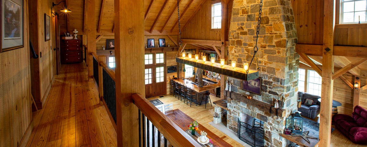 A view from a wooden loft in a luxury home overlooking a spacious rustic living area with a stone fireplace, dining table, chandelier, and cozy seating, all featuring exposed wood beams and natural light.