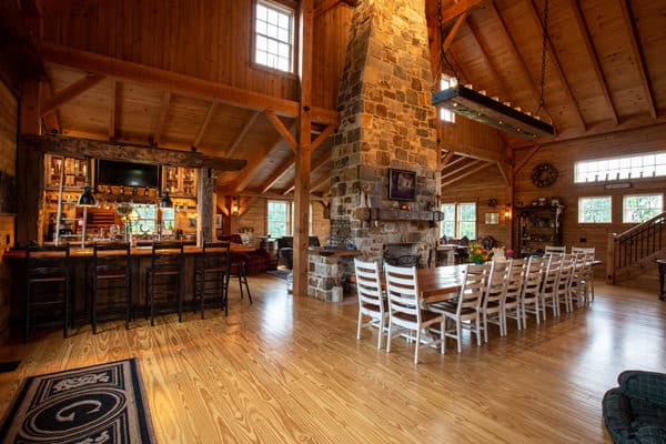 Spacious rustic cabin interior with high wooden ceilings, a large stone fireplace, and a long dining table with white chairs—evoking the charm of a luxury horse barn. Cozy bar area with wooden stools on the left. Natural light streams through multiple windows.