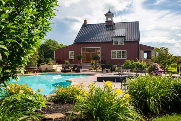 A red party barn-style house with a black metal roof stands behind a landscaped backyard featuring a swimming pool, colorful flowers, green plants, and patio seating under a partly cloudy sky.