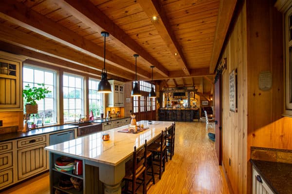 A spacious rustic kitchen with wood-paneled walls and ceiling, a large island with chairs, pendant lights, white cabinets, and large windows letting in natural light—perfect for entertaining in a luxury home or transforming into a stylish party barn.