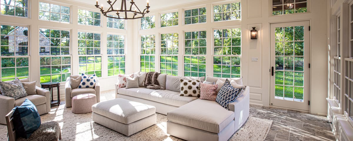 Bright sunroom with large windows, a white sectional sofa, patterned pillows, armchairs, a central ottoman, light rug, chandelier, and views of a lush green backyard—perfect for relaxing in this luxury home inspired by classic bank barn design.