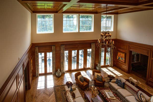 Spacious living room with high ceilings, large windows, wooden walls and ceiling—this luxury home features patterned rugs, several couches, and armchairs. Sunlight streams in through tall windows and glass doors, brightening the elegant space.