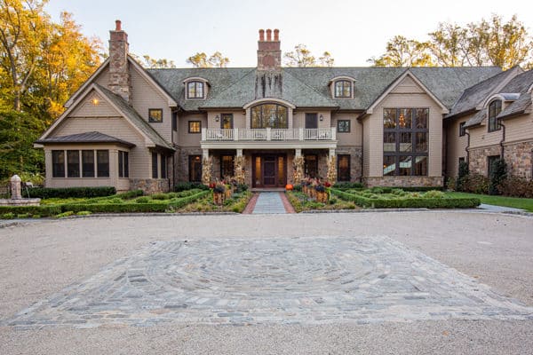 Large, elegant luxury home with stone and wood exterior, gabled roof, multiple chimneys, and large windows; landscaped front yard with walkway, shrubs, and pumpkins near the entrance evokes classic bank barn charm.