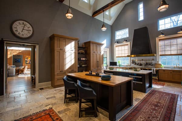 Spacious luxury home kitchen with high ceilings, wooden cabinets, a large island with two chairs, hanging lights, and tall windows. A clock hangs on the wall while sunlight streams in from the windows.