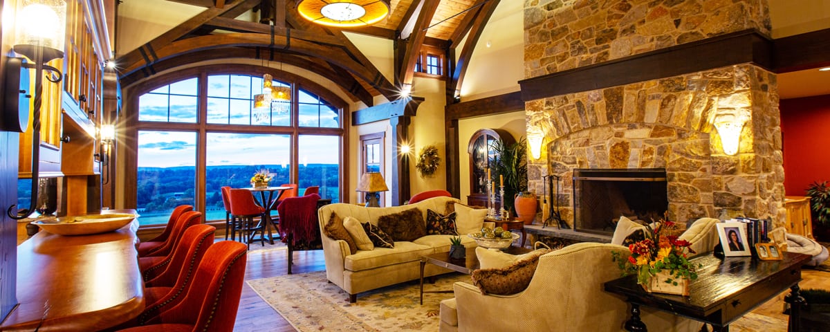 Cozy living room in a luxury home with a large stone fireplace, plush sofas, and wooden beams. Tall arched windows reveal scenic outdoor views. Warm lighting enhances the inviting, rustic yet elegant atmosphere.