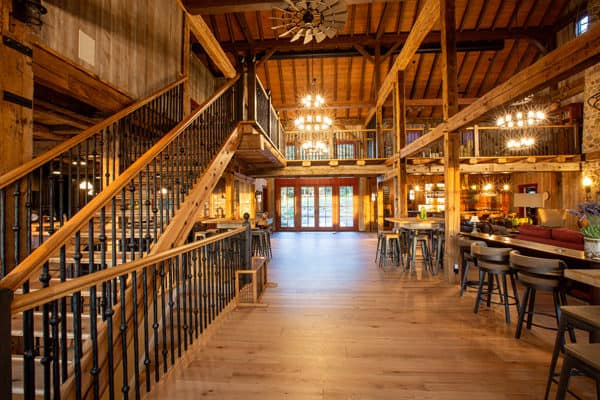 Spacious, rustic interior of a luxury horse barn-style restaurant with wooden beams, high ceilings, chandeliers, a staircase, and modern seating. Warm lighting creates a cozy and inviting atmosphere perfect for gatherings.