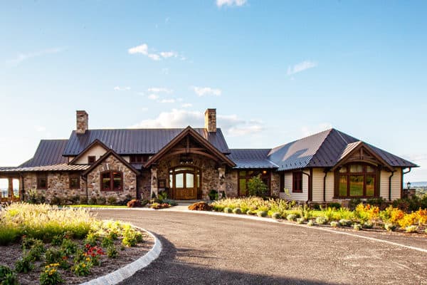 A large, rustic-style luxury home with a stone and wood exterior, arched entryway, and landscaped circular driveway, surrounded by gardens under a clear blue sky.