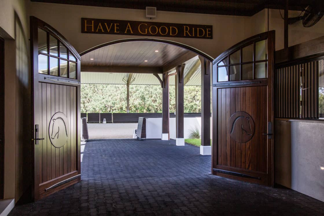 Open wooden barn doors with horse engravings lead from this luxury horse barn to an outdoor riding arena. Above the doors, a sign reads HAVE A GOOD RIDE. Trees and a covered area are visible in the background.