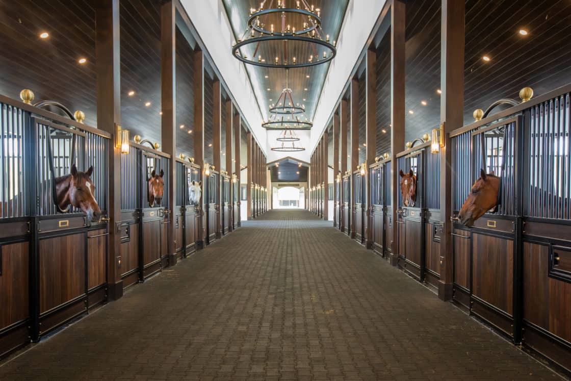 A wide, well-lit luxury horse barn with elegant chandeliers, dark wood paneling, and several horses looking out from their individual stalls along both sides of a central brick pathway.