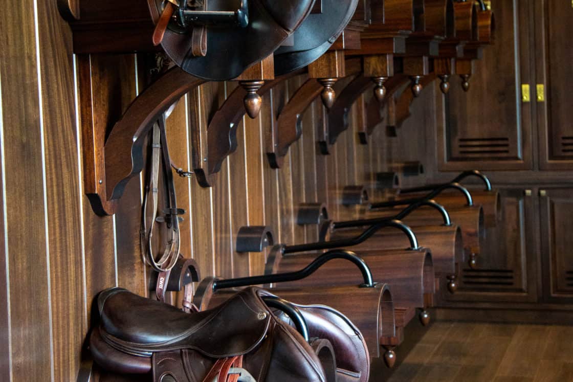 Several brown leather horse saddles are neatly hung on wooden racks along a wood-paneled wall in a luxury horse barn tack room with dark wood flooring and cabinets.
