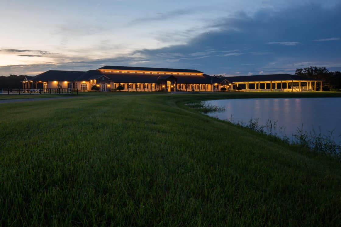 A luxury home with glowing lights sits beside a pond at dusk, surrounded by lush green grass and a clear sky transitioning from day to night.