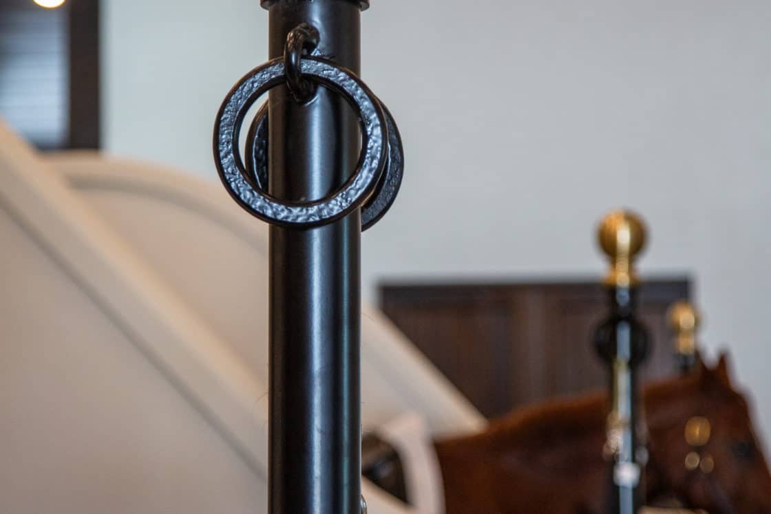 A close-up of a black metal stanchion with a gold ball top and two black rings, set indoors near a white divider in a luxury horse barn, with another similar stanchion and a brown horse in the background.