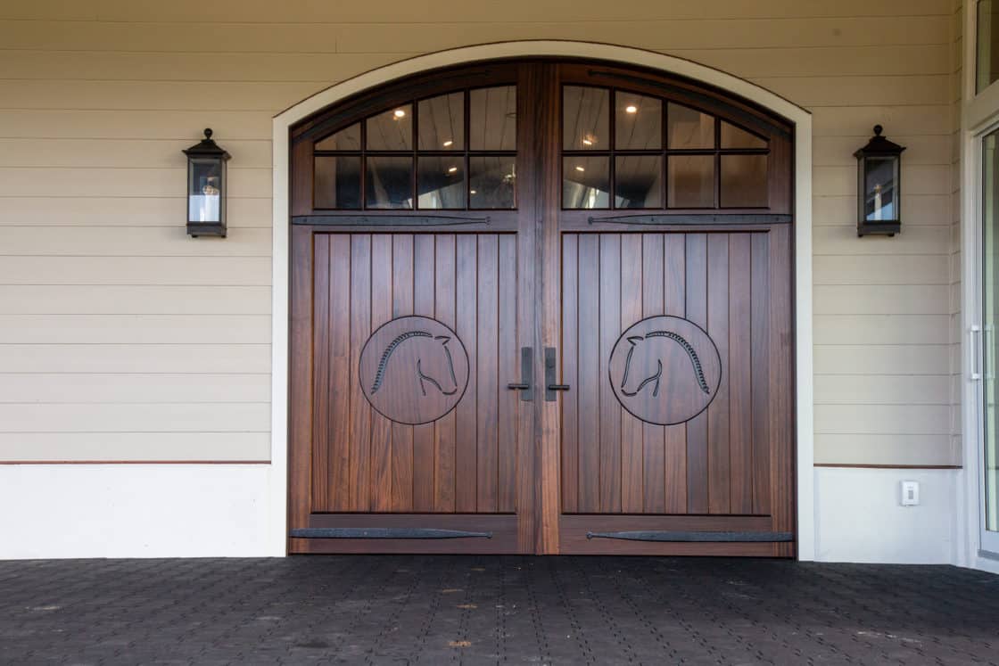 A pair of large wooden double doors with horse head carvings in circular frames, set in a light-colored wall with two black lantern sconces on either side, evokes the elegance of a luxury horse barn. Arched windows crown the entry.