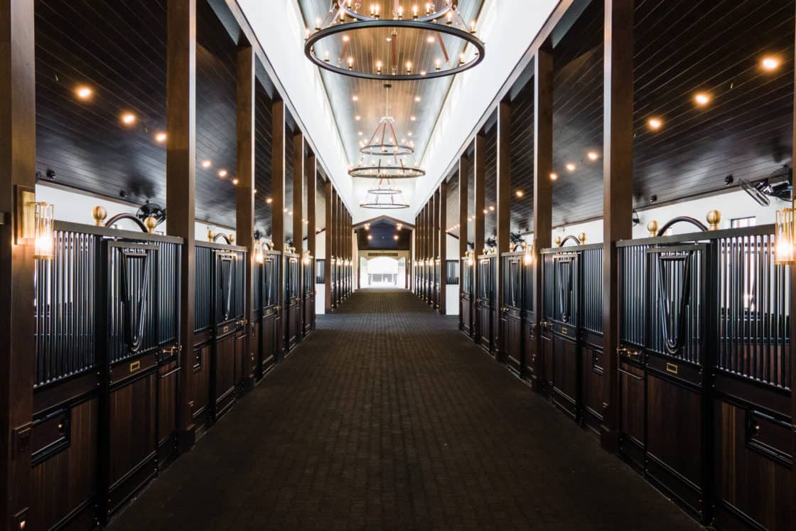 A spacious, modern luxury horse barn with dark wood stalls and black metal bars, illuminated by elegant circular chandeliers and ceiling lights along a wide central aisle.