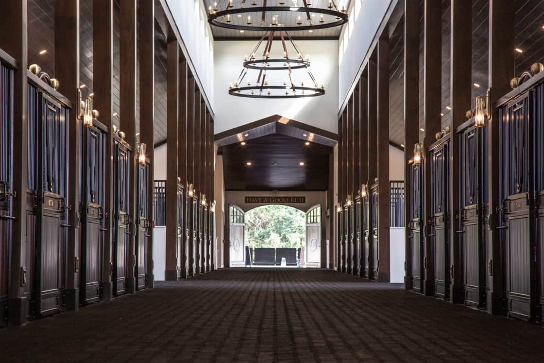 A wide, clean luxury horse barn hallway with dark wooden stalls, high ceilings, chandeliers, and sunlight streaming in from the open doors at the far end—perfect for a party barn or elegant luxury home setting.