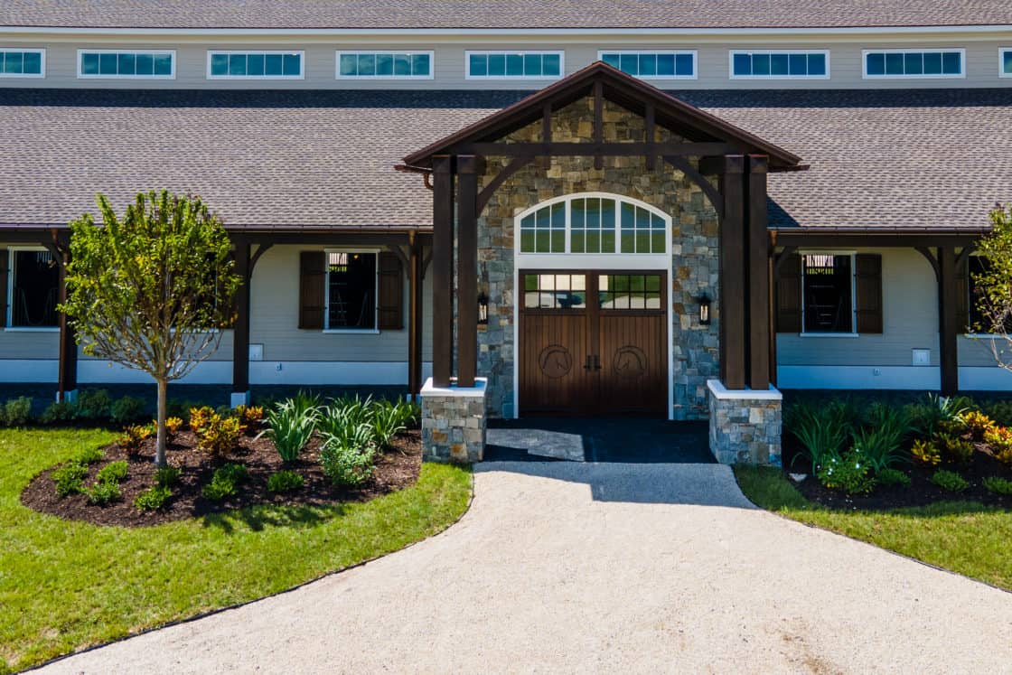 A stone and wood entrance of a large, modern bank barn with double wooden doors, landscaped garden beds, green grass, and a gravel driveway leading to the entrance—perfect for a luxury home or party barn setting.