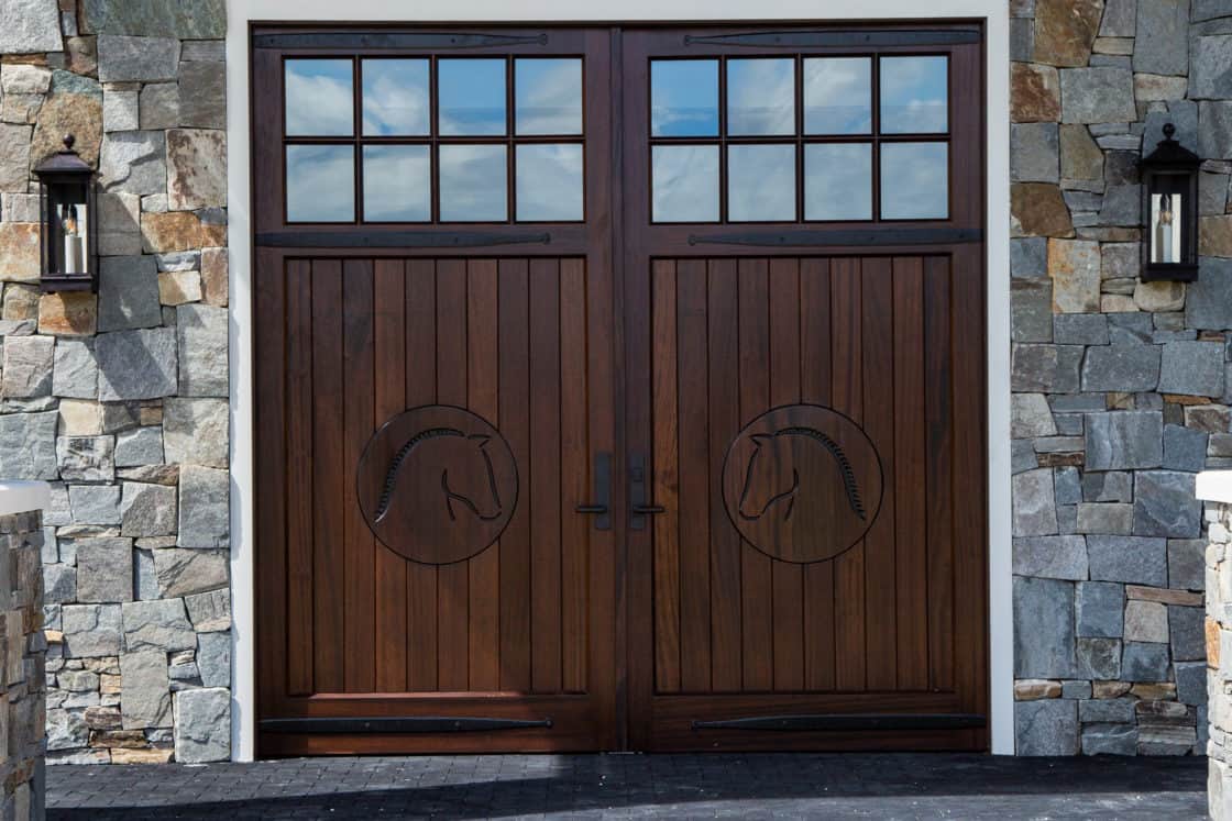 A large, double wooden door with square glass panels and horse head carvings, set in a stone bank barn with an arched window above and two black wall lanterns on either side.