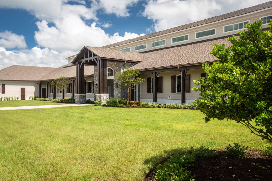 A modern ranch-style party barn with a covered entrance, tan siding, and brown roof, surrounded by green grass, bushes, and trees under a partly cloudy blue sky.