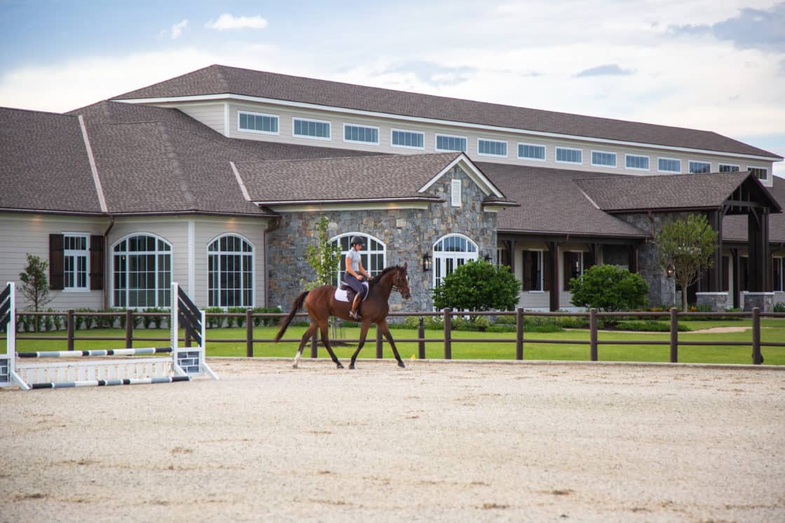 A person rides a brown horse in an outdoor arena near a luxury horse barn with stone and siding, surrounded by green landscaping and a wooden fence.