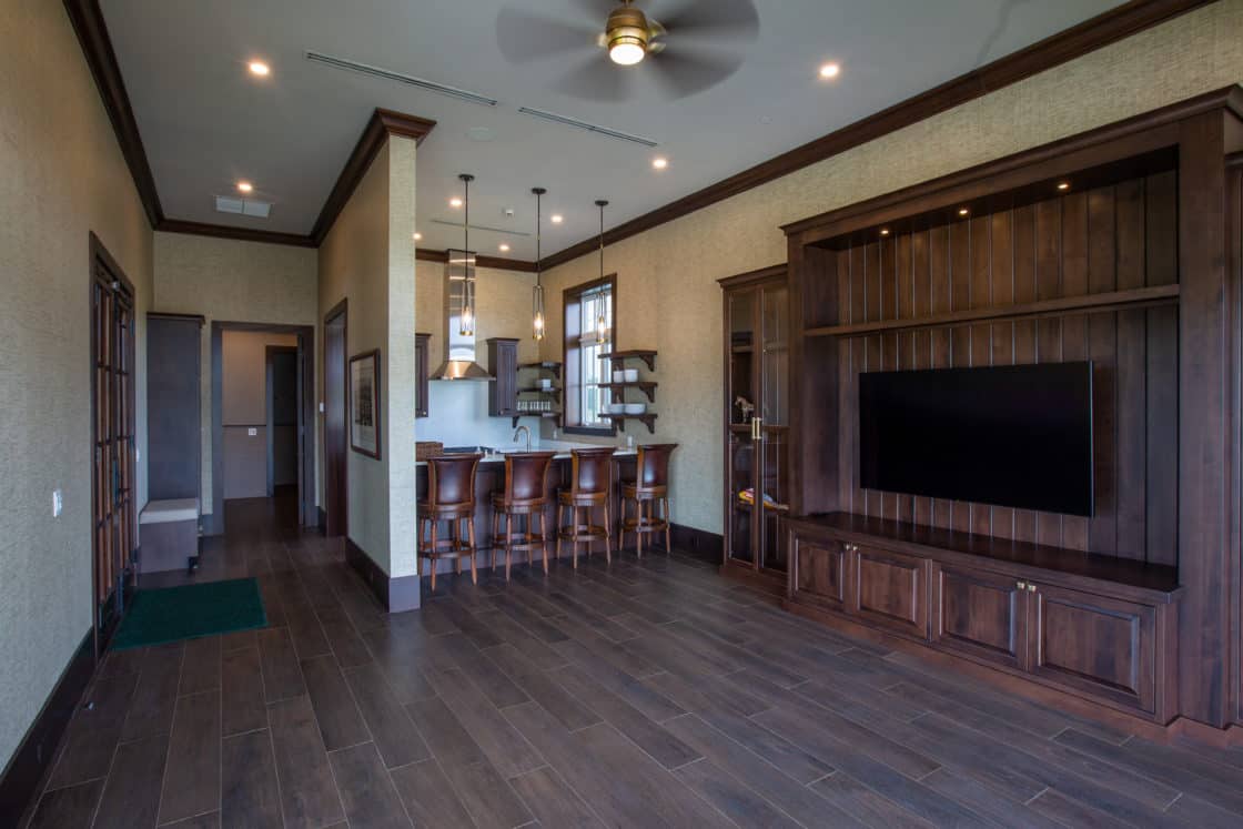 A modern living space with dark wood flooring, a built-in entertainment center, and a bar area evokes the relaxed elegance of a luxury horse barn, featuring four chairs, pendant lights, open shelving, neutral walls, and ceiling fans.