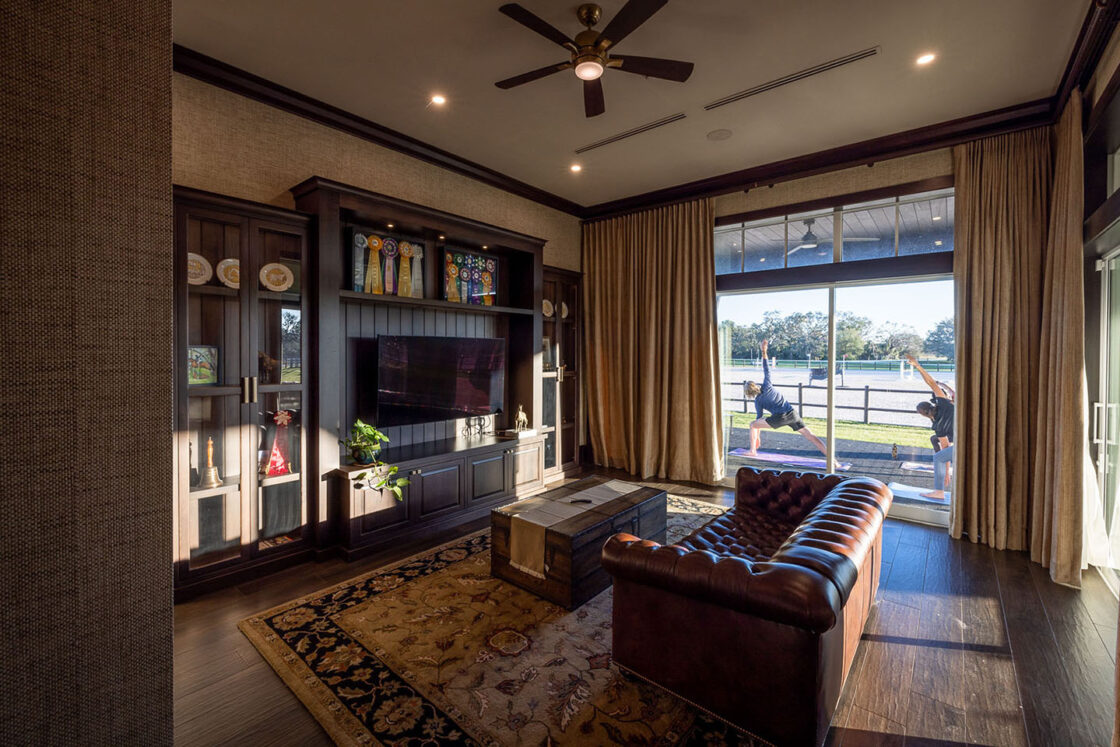 Cozy living room with a leather sofa, patterned rug, wooden coffee table, and glass-front cabinets displaying ribbons and trophies. Large window reveals a luxury horse barn with horses and riders outside. Warm lighting and ceiling fan above.
