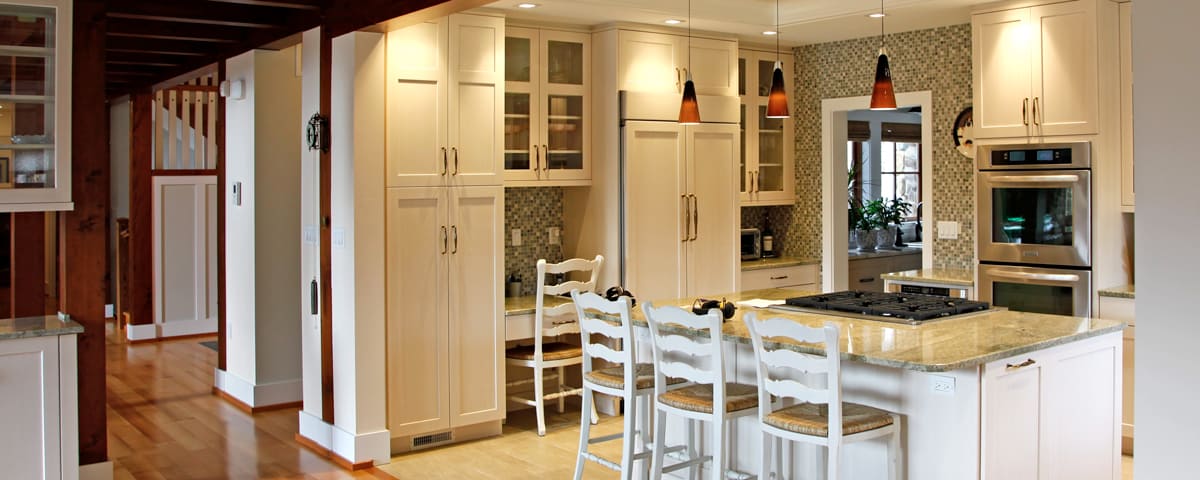 Bright, modern kitchen in a luxury home features cream cabinets, a central island with three white chairs, built-in stove, double ovens, pendant lights, mosaic tile backsplash, and hardwood floors. Natural light streams in through the window.