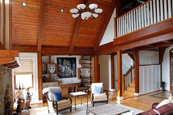 A spacious, rustic living room with a high, wooden vaulted ceiling reminiscent of a party barn, exposed beams, a stone fireplace, two armchairs, built-in shelves, and a staircase with a white railing leading to an upper loft area.