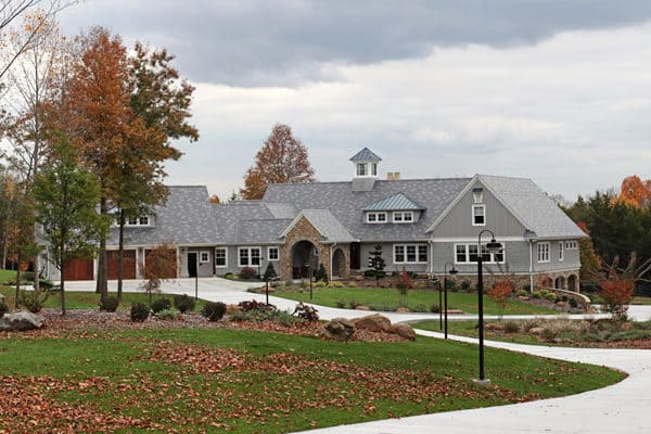 A large, gray luxury home with a stone arch entrance and cupola sits on a curved driveway, surrounded by autumn trees and a lawn covered with fallen leaves under a cloudy sky.