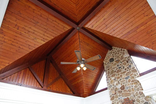 A vaulted wooden ceiling with triangular panels, a ceiling fan with lights in the center, and a stone fireplace extending up to the ceiling create the warm ambiance of a luxury home.