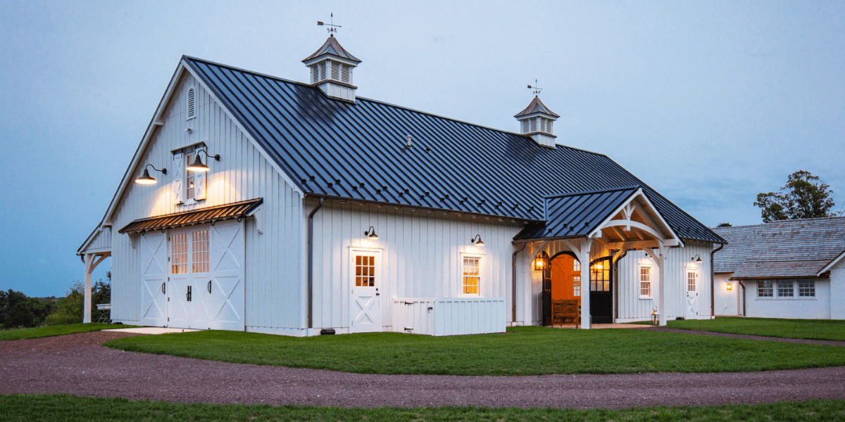 A dark blue bank barn with a tin roof stands on a grassy hill at sunset, surrounded by green trees and a partially cloudy sky with warm orange and yellow tones.