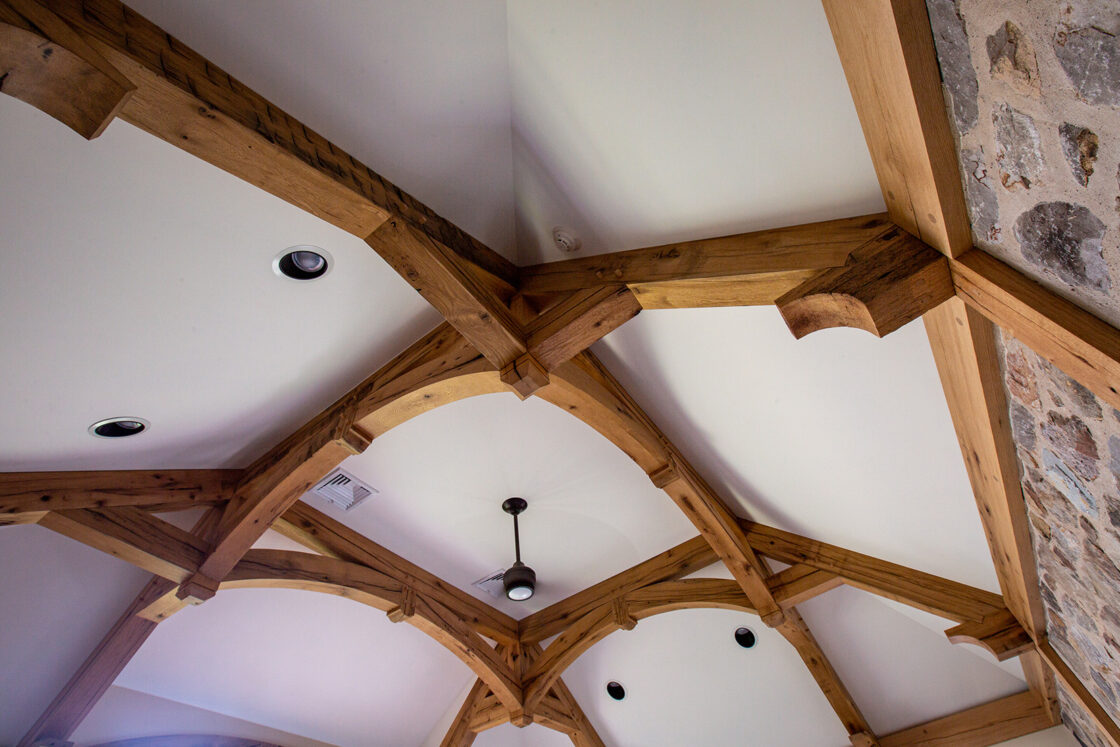 Exposed wooden beams form an intricate pattern on a white vaulted ceiling in this luxury home, featuring recessed lights, a central ceiling fan, and a partial stone wall visible on the right side.