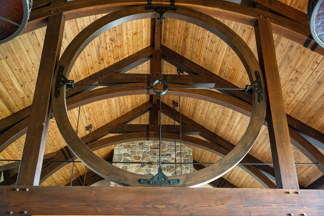 Wooden beams and a large circular wooden structure are set against a vaulted wood-paneled ceiling, with a stone wall in the background. This luxury horse barn image showcases remarkable architectural craftsmanship.