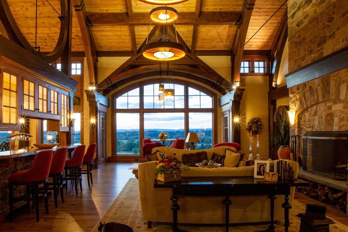 Spacious living room with high wooden ceilings, large windows overlooking a scenic view, a stone fireplace, warm lighting, and wood floors—this luxury home features a beige sofa, red barstools at the kitchen counter, and a cat lounging nearby.