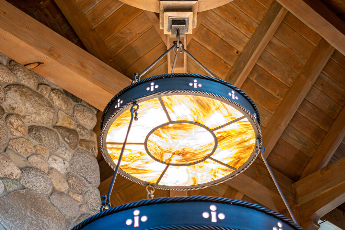 Two large, circular stained glass chandeliers with warm yellow lights hang from a wooden ceiling with exposed beams and a stone wall, evoking the refined ambiance of a luxury home or expertly crafted bank barn.