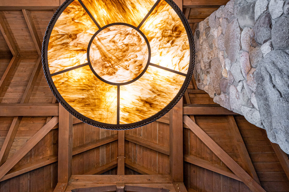 A large, circular amber light fixture hangs from a wooden vaulted ceiling in this luxury horse barn, next to a tall stone column, creating a warm and rustic atmosphere.