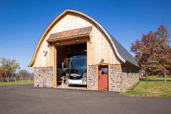 A large wooden and stone bank barn with a curved roof houses a modern RV inside. The barn sits on a paved driveway, surrounded by grass and trees under a clear blue sky.