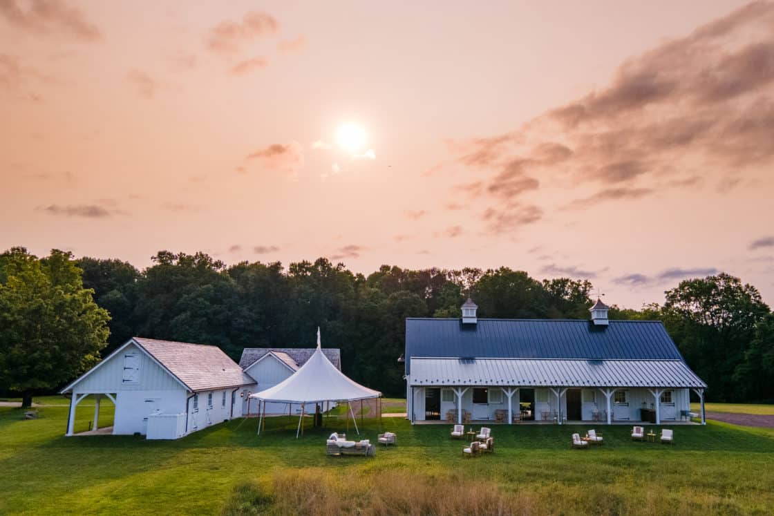 landscape view of white party barn with white tent and green grass in front of trees