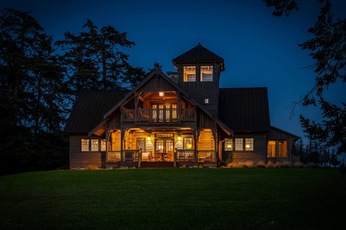 A large, rustic luxury home with multiple balconies and a tower, warmly lit from within, stands surrounded by trees and grass against a deep blue evening sky.