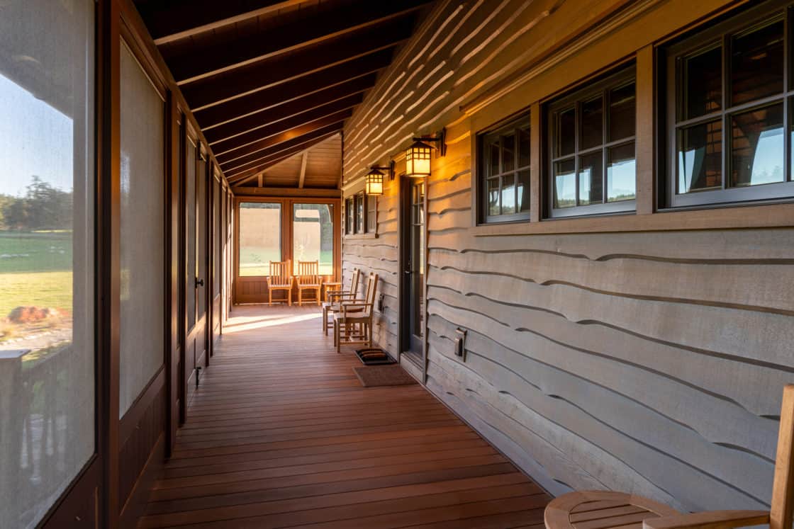 A covered wooden porch with a wavy-textured wall, windows, warm wall lanterns, and two wooden chairs evokes the charm of a luxury horse barn, with a view of more chairs at the far end overlooking a grassy yard.