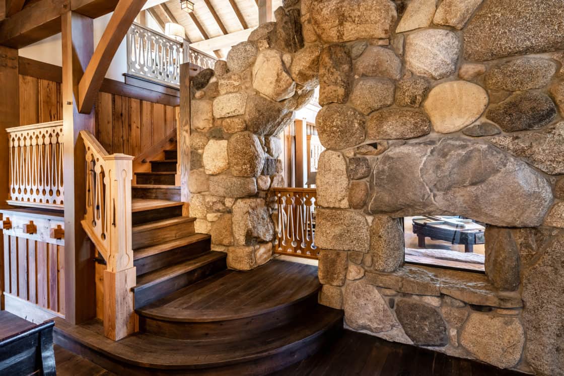 A rustic interior, reminiscent of a luxury horse barn, features a curved wooden staircase with ornate railings and a large stone wall, creating a cozy and inviting atmosphere with warm natural wood and stone textures.