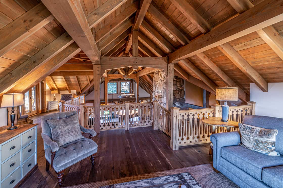 Cozy loft with rustic wooden beams, vaulted ceiling, stone accents, armchair, and side table with lamp—just like a luxury horse barn. Warm lighting and a railing overlooking the lower level create an inviting atmosphere.