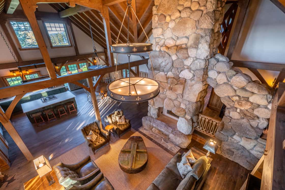 A rustic living room with exposed wooden beams reminiscent of a bank barn, a large stone fireplace, circular chandeliers, cozy seating, and an open view of the kitchen and dining area, all bathed in natural light.