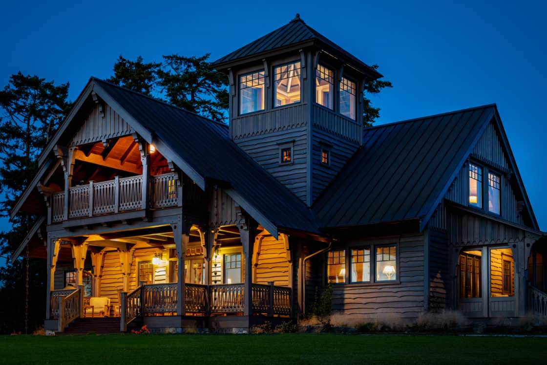 A warmly lit luxury home with a covered porch and an upper-level tower stands against a deep blue evening sky, surrounded by trees and a green lawn.