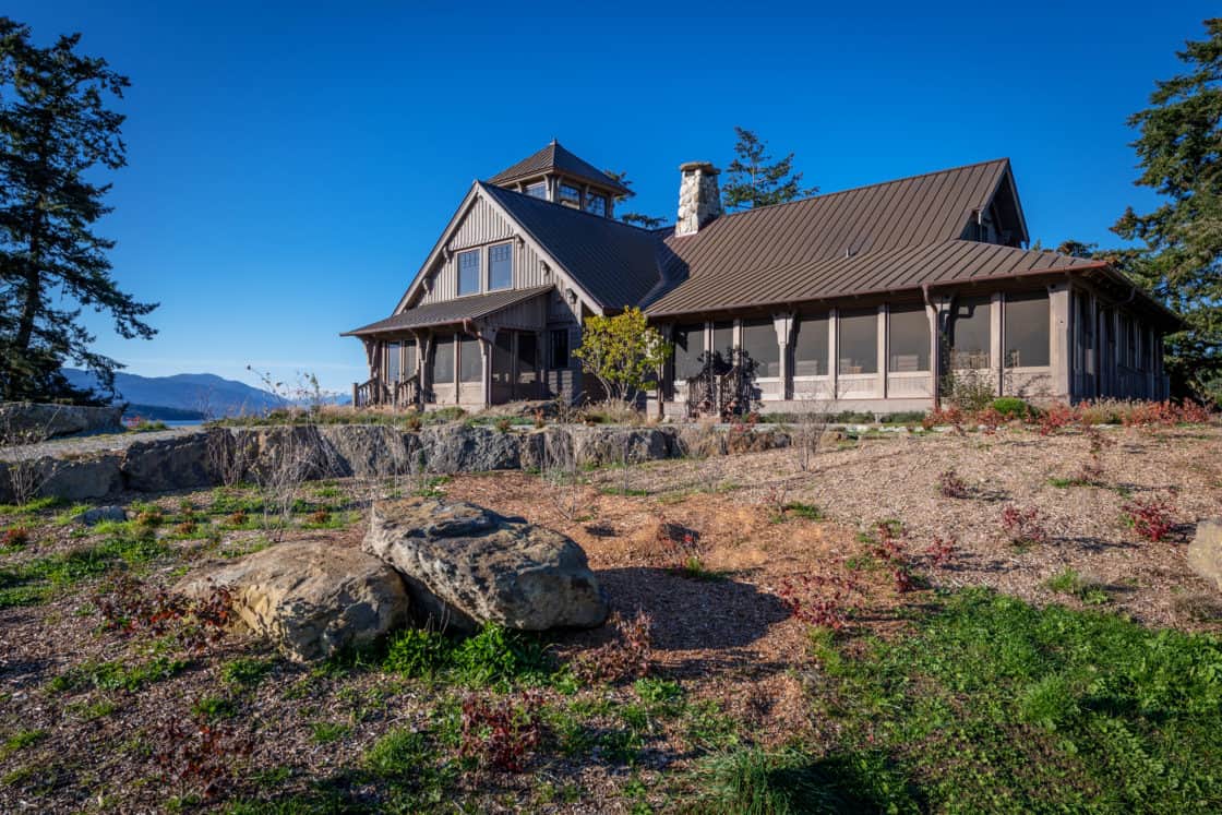 A rustic house with a wraparound porch sits on a rocky, landscaped hill under a clear blue sky, surrounded by sparse greenery and trees—a charming scene reminiscent of a bank barn or future luxury home.
