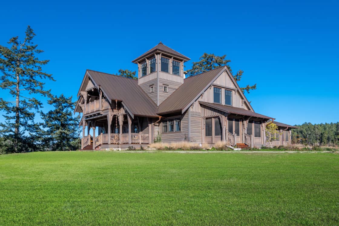 A large, rustic wooden house with a tower and wraparound porch sits on a green lawn, surrounded by trees under a clear blue sky, reminiscent of a classic party barn retreat.
