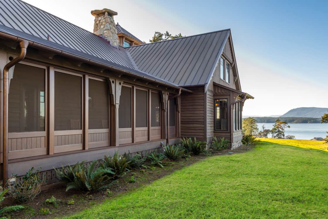 A brown wooden house with a metal roof and screened porch sits on a grassy lawn overlooking a calm body of water, with distant hills and a classic bank barn under a clear sky.