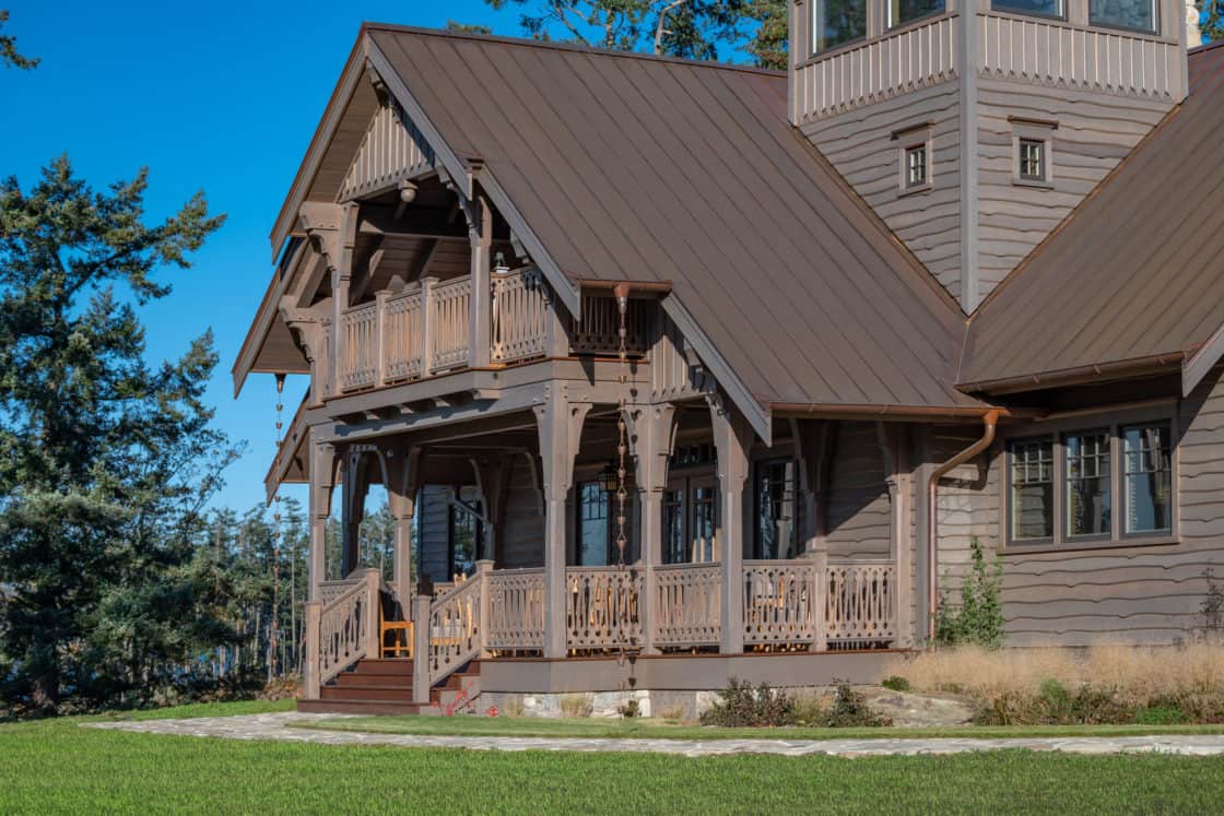 A large wooden luxury home with a metal roof, multiple balconies and porches, surrounded by green grass and trees under a clear blue sky.