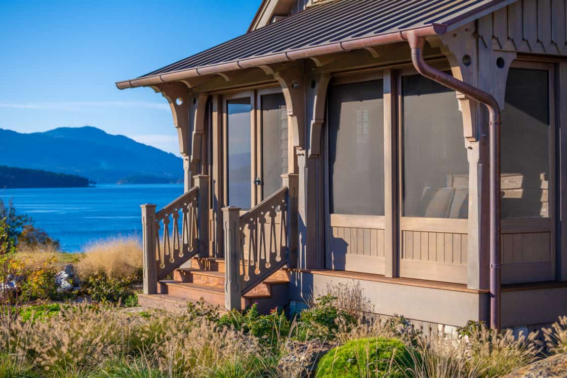 A cozy wooden cottage with screened porch and decorative railings overlooks a calm blue lake and distant mountains under a clear sky, offering the charm of a luxury home on a sunny day.